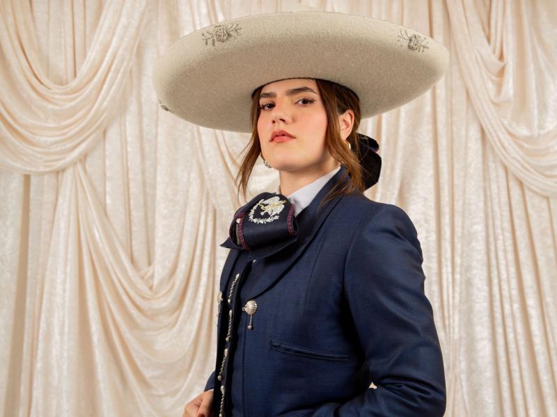 Woman posing in navy blue traje de charro outfit with a cream colored hat