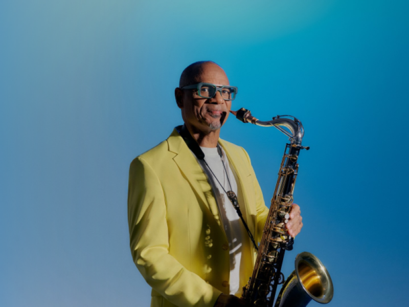African American man wearing a yellow blazer holding a saxophone.
