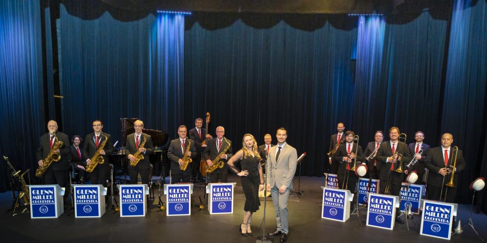 Formal photo of the Glenn Miller Orchestra on stage with the lead female singer and male band director