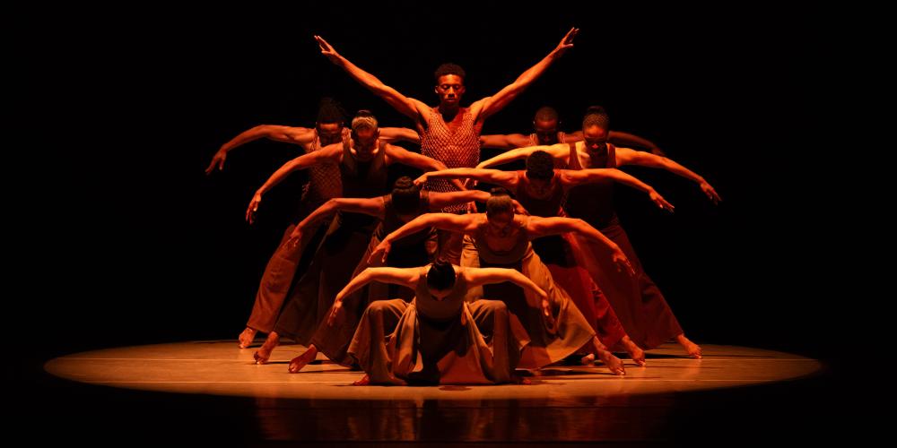 Group of 9 dancers under a spotlight with arms outstretched wide