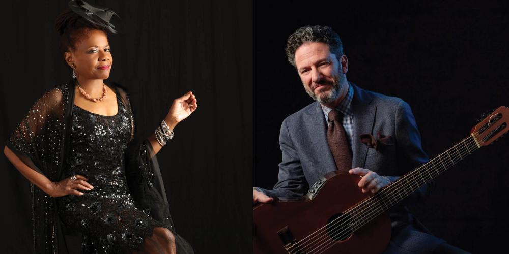 A woman in a black dress sits on a chair, posing with a smile, and a man in a gray suit sits with his guitar