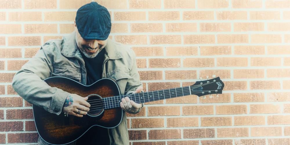 Blaine Long, sporting a beard and wearing a driver cap and khaki jacket leans against a red brick wall with his amber acoustic guitar in his hands