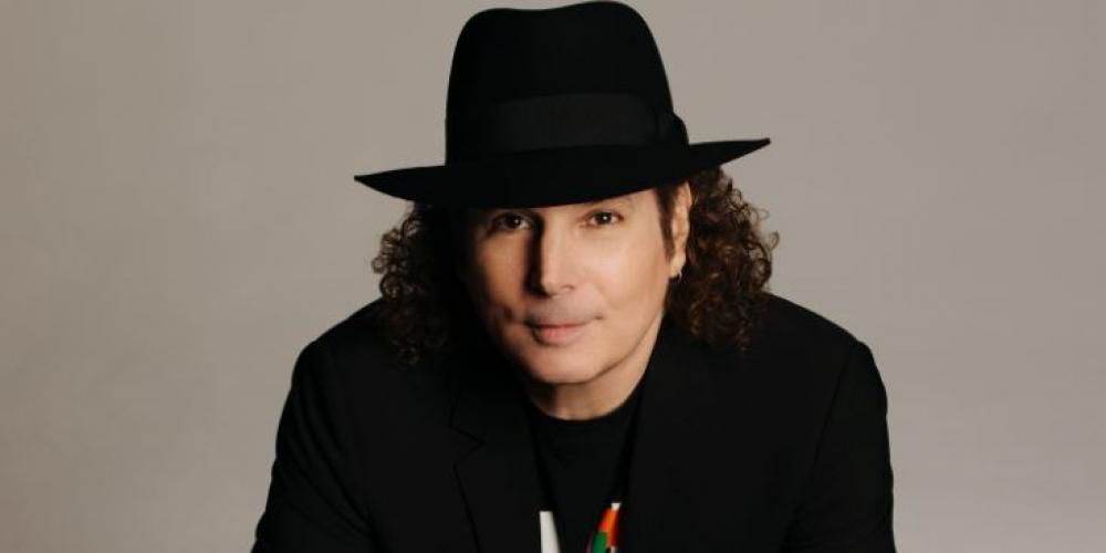 A man with shoulder length curly hair leans over a counter, a saxophone under his hands. He wears all black and a black fedora, smiling slyly at the camera