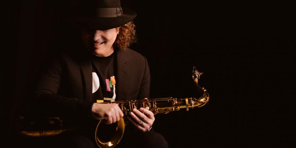 A man with shoulder length curly hair wears all black, blending in with the black background so that you only see his hands holding a saxophone and his face smiling under his fedora.