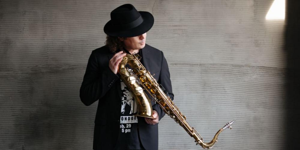 A man with shoulder length curly hair stands against a gray concrete wall, dressed in black with a black fedora. He holds his saxphone in his hands at an angle 