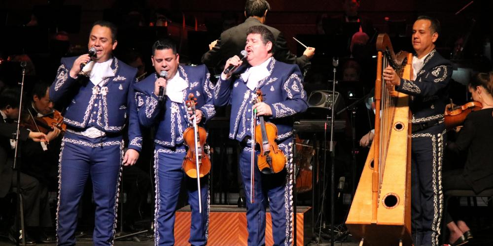 Four men wear blue mariachi suits with silver buckle accents. Two hold violins, one plays a harp and the other sings