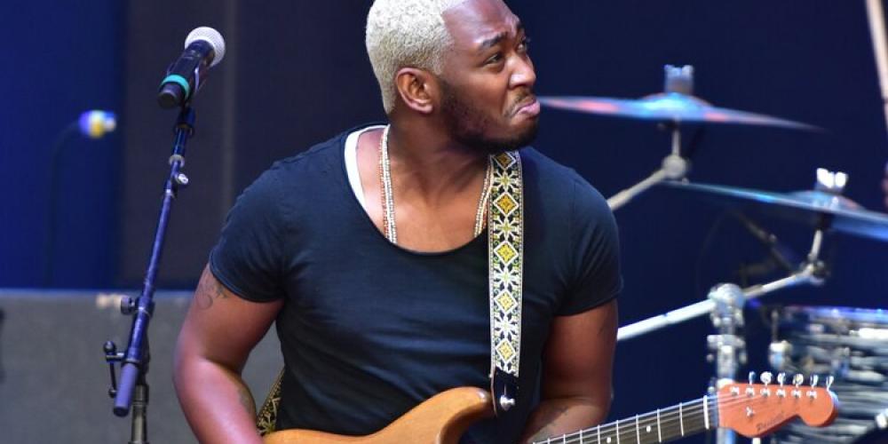 A man plays guitar on stage, his guitar is golden, he is wearing all black and he has tight curly hair colored platinum blond