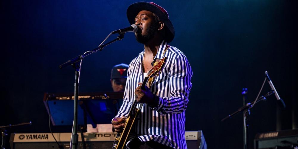 A man stands on a stage behind a microphone, singing, eyes closed. He is wearing a black and white striped button up shirt and a brown fedora. A guitar is slung low on his hip as he strums. 