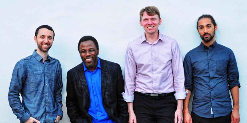 Four men stand against a white wall smiling at the camera. Three wear blue button-up shirts and one wears a pink button-upshirt. 