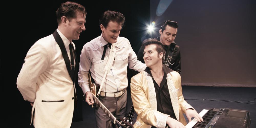 Four men dressed in suits are gathered around a piano with two holding guitars