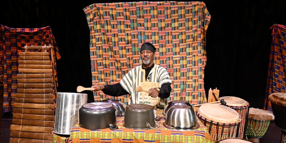 Percussionist Keith Johnson sits in traditional African clothing, playing a variety of percussion instruments including African drums, pots and pans.