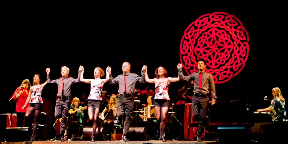 Irish step dancers hold hands while a band plays behind them, a celtic knot illuminated behind them