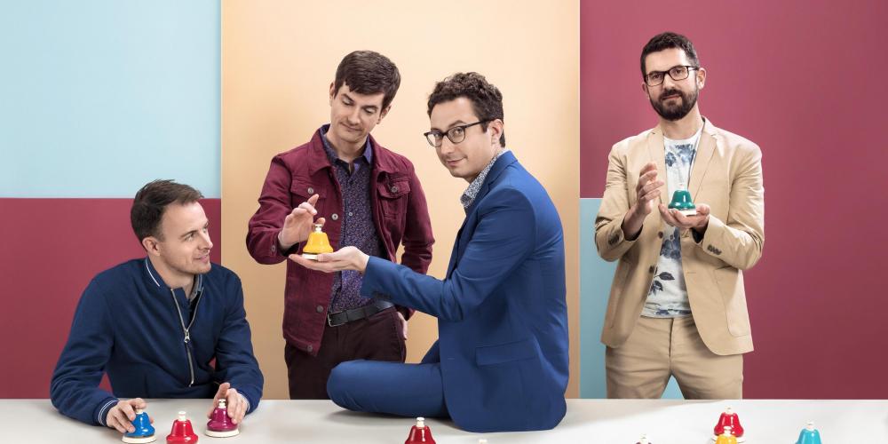 Four men dressed in colorful clothing standing in front of a colorful block background are in front a table with bells of different colors