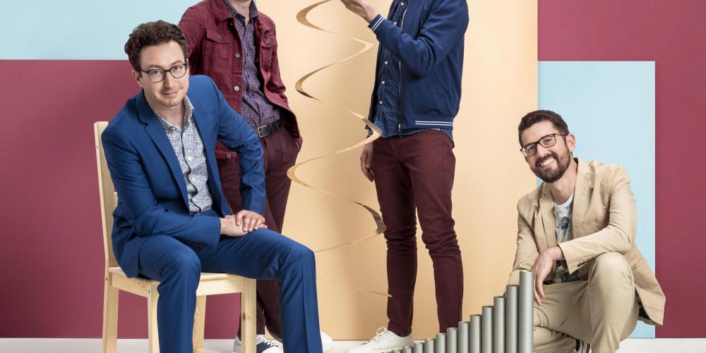 Four men in colorful clothing with a colorful block background standing with two percussion instruments - one is tall and swirled brass and the other is silver tubes