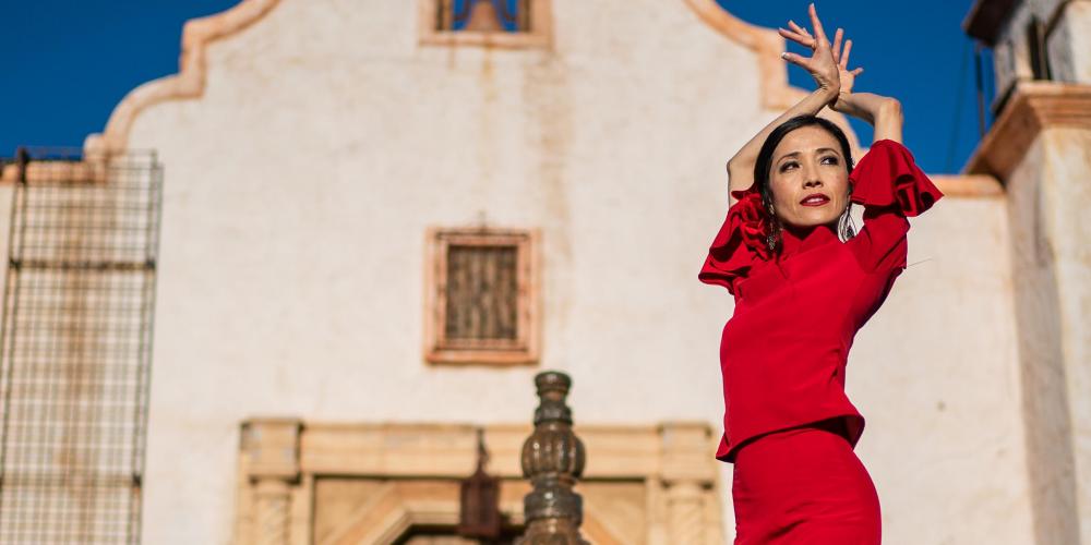A tall woman poses in front of a mission in a red dress with ruffles at the hem