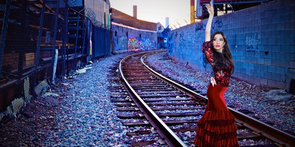 A tall woman poses by railroad tracks in a red velvet dress with layers of ruffles from her knees to the ground