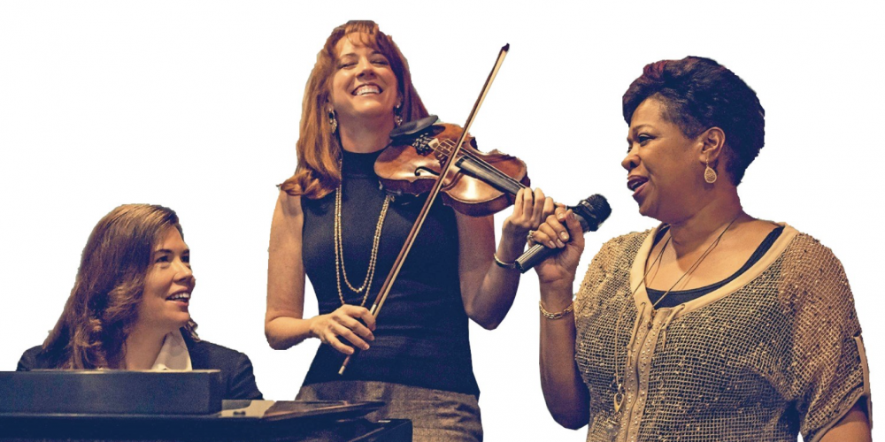 Three women are in a half circle; one plays piano, one plays violin and one sings 