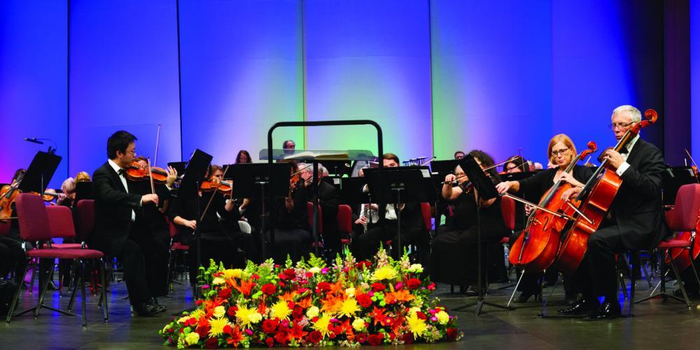 Photo of orchesra with flowers on the floor behind conductor.