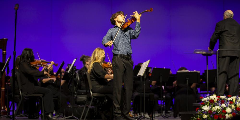 A young man plays the violin on stage with an orchestra behind him
