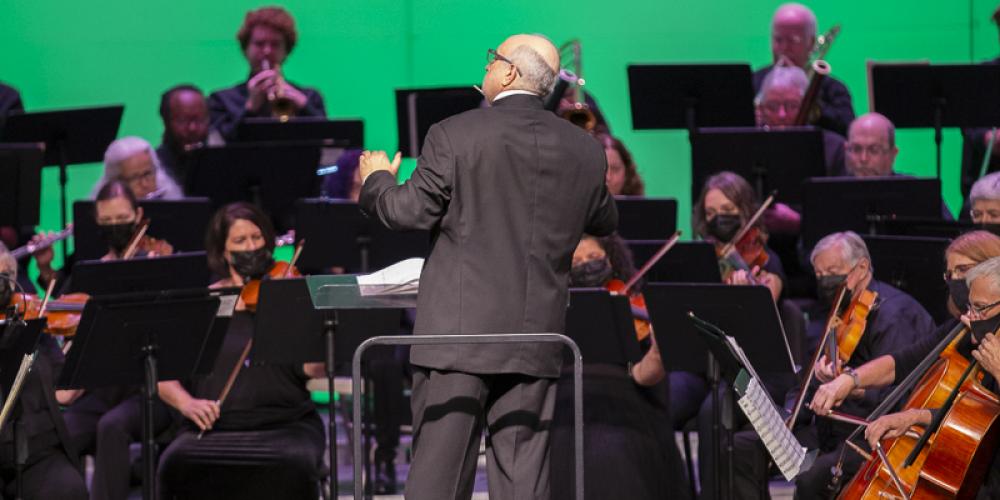 Musicians in a symphony play while the conductor stands in front of them, his back to the camera
