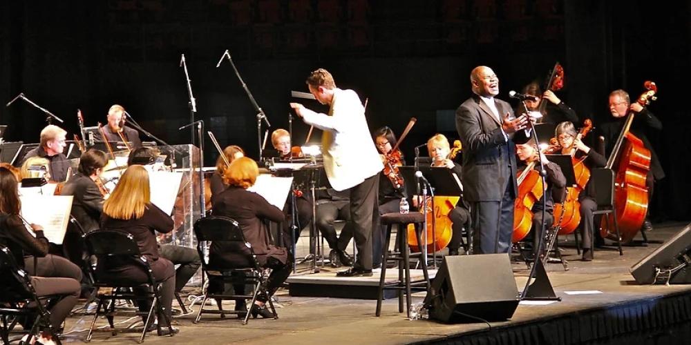 A man dressed in a black suit sings into a microphone on a stage with an orchestra behind him