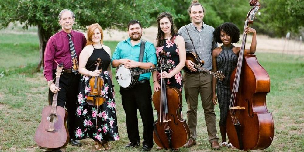 A group of six people stand in a field, holding their string instruments