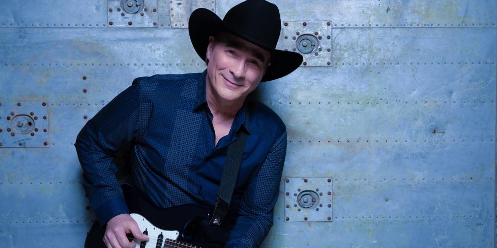 A man in a dark blue shirt stands against a blue wall, electric guitar in hand and a black hat on his head