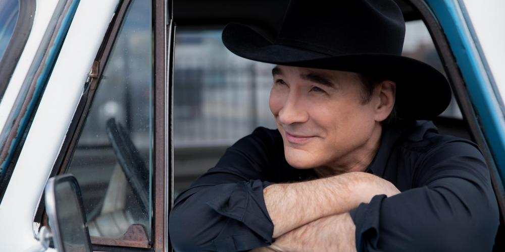 A man leans out the window of a baby blue truck, arms folded and his chin resting on his forearm. He wears a black hat and shirt. 
