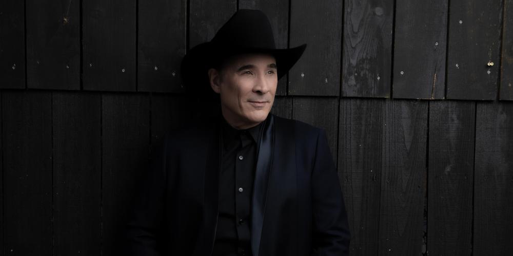 A man all dressed in black - hat, shirt and jeans - leans against a dark brown wall covered in wood slats