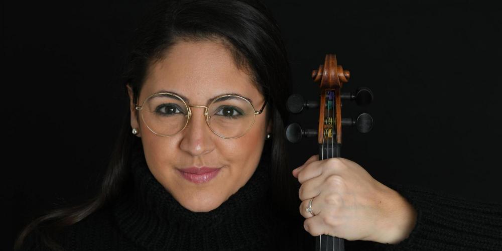 A woman with long dark hair pulled back from her face wears glasses and a black turtleneck sweater. She holds a violin in her left hand. 