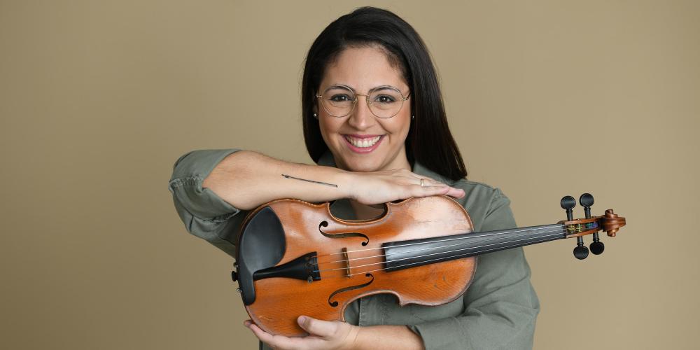 A woman with long black hair and glasses faces the camera holding her violin between her arms