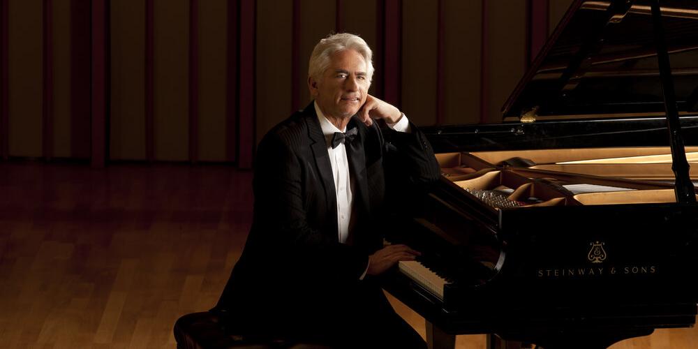 David Benoit - a man with silver hair - sits at a grand piano in a tuxedo looking at the viewer with a smile
