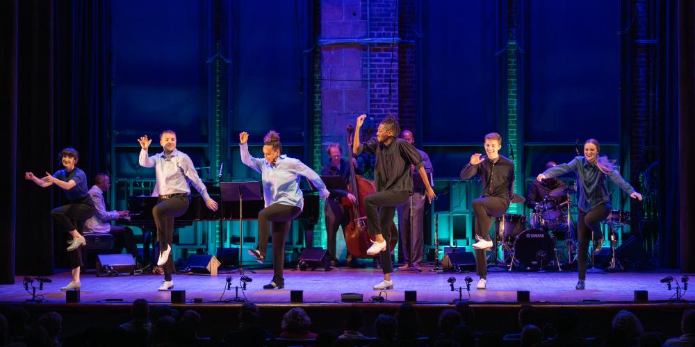 six dancers on stage smiling with right arms up and left knees raised up