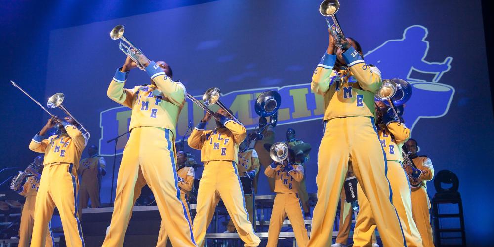 Horn line from Drumline Live wearing yellow uniforms in front of a blue backdrop.