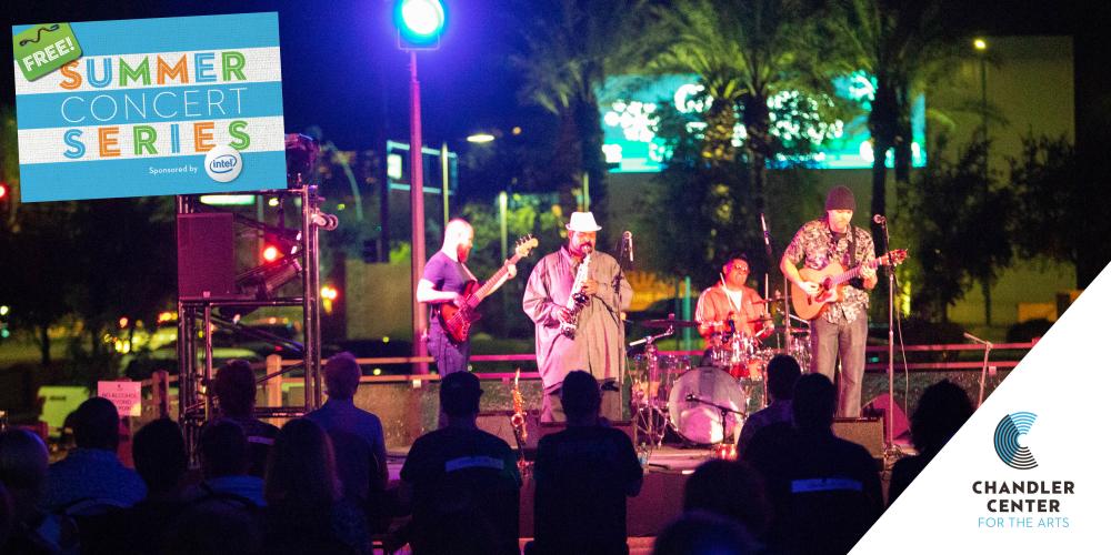 Four men play music on a stage that is outside. It is dark out and lights of the city are behind them. Two play guitar, one plays drums and the lead man plays a saxophone.