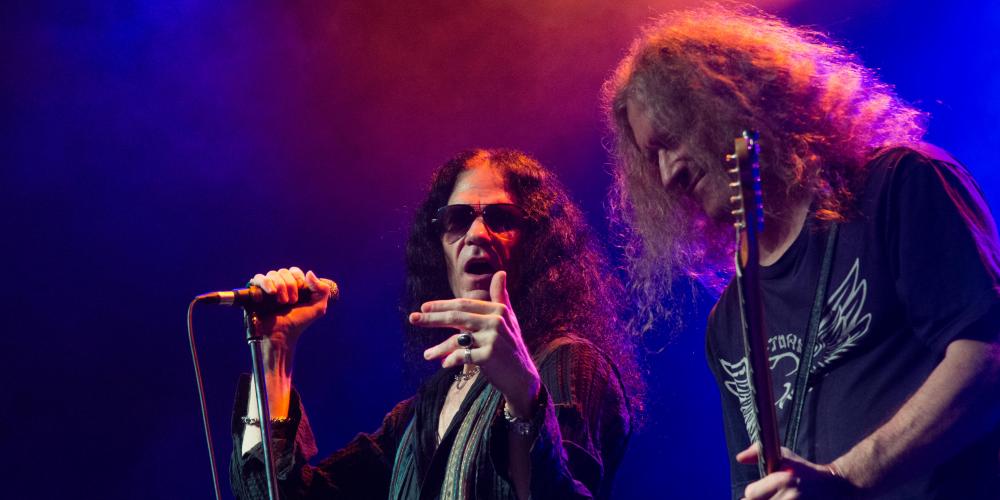A man with long hair and sunglasses, sings into a microphone while a man with long hair plays a guitar next to him