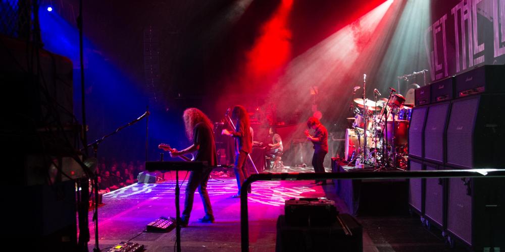 The image is a back stage view of a rock band playing with colorful lights overhead - a guitarist and drummer are in the foreground