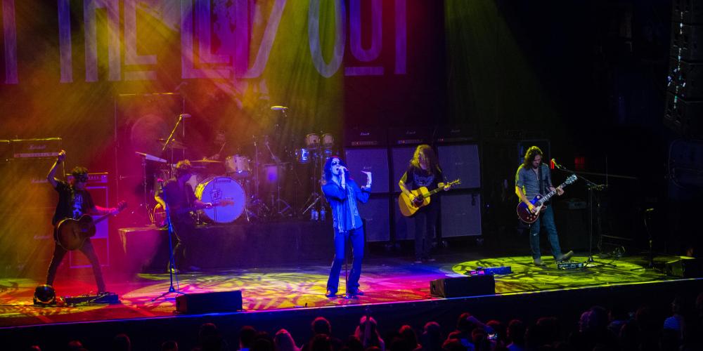 Wide shot of a stage with pink and yellow lights overhead, three guitarists, a singer and drummer are on stage