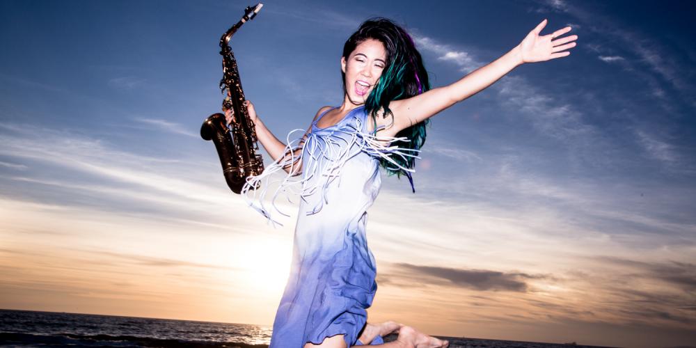 A woman with long black hair is jumping in the air, saxophone in hand, at the beach with a sunset behind her 