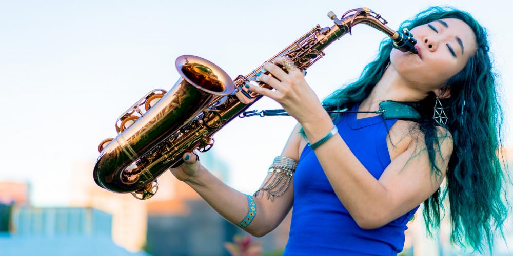 A woman with long wavy green hair, wearing a royal blue shirt, plays a saxophone with eyes closed and her head leaning back