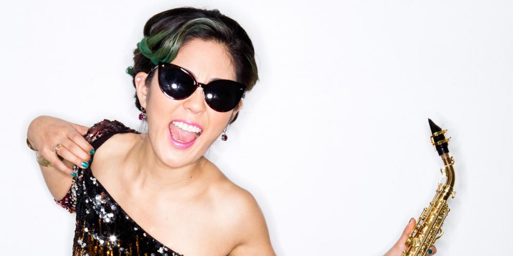 A woman with an updo wears sunglasses and a sequin dress, smiling and holding a saxophone