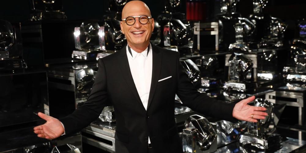 A white man with no hair standing on stage wearing a black suit and white shirt and glasses