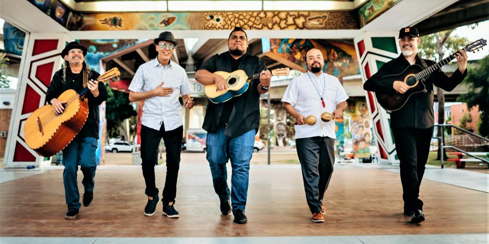 Five men walk through a courtyard toward the camera, four hold instruments