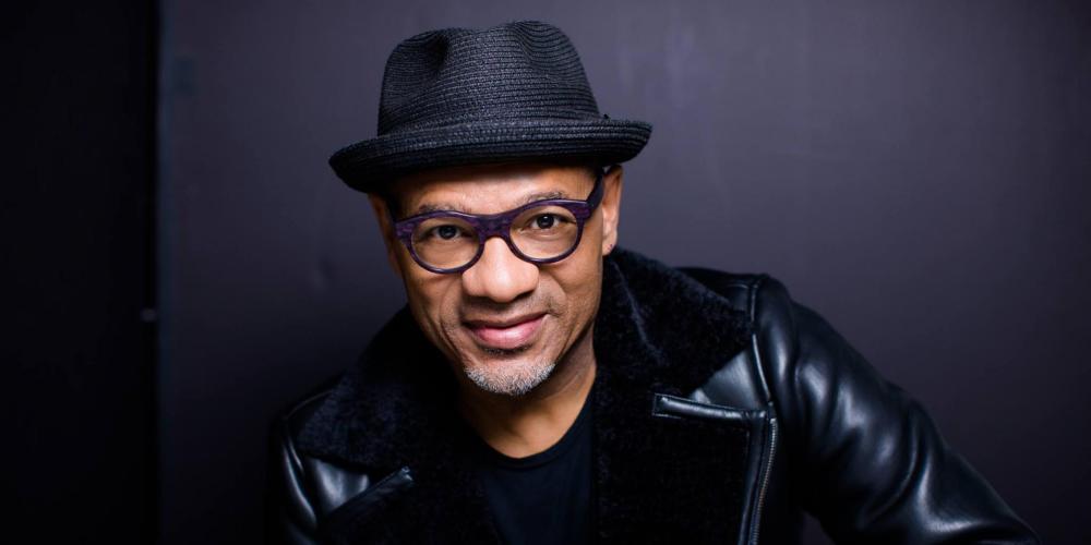 A black man wearing a black leather jacket, glasses and a black fedora looks at the camera.