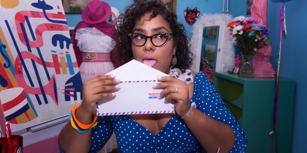 La Marisoul Sitting at a desk in a colorful blue dress licking an envelope to seal it