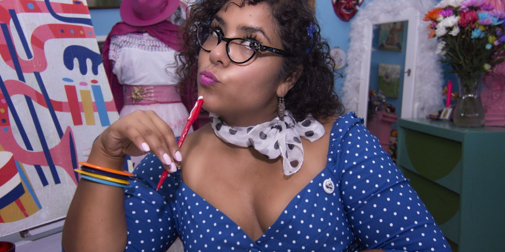 La Marisoul sitting at a desk in a colorful blue dress writing a letter and looking off to the side with a silly look on her face