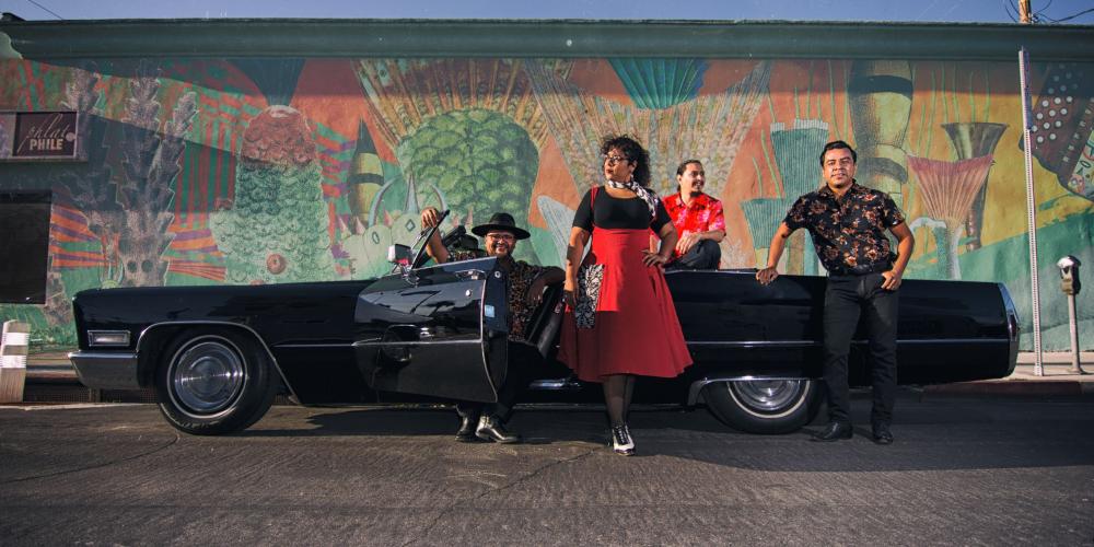 La Santa Cecilia group standing around a classic black Cadillac convertible