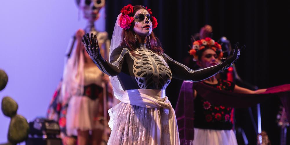 two performers on stage with one in focus holding arms out and dressed as a dia de los muertos skeleton with florals in hair