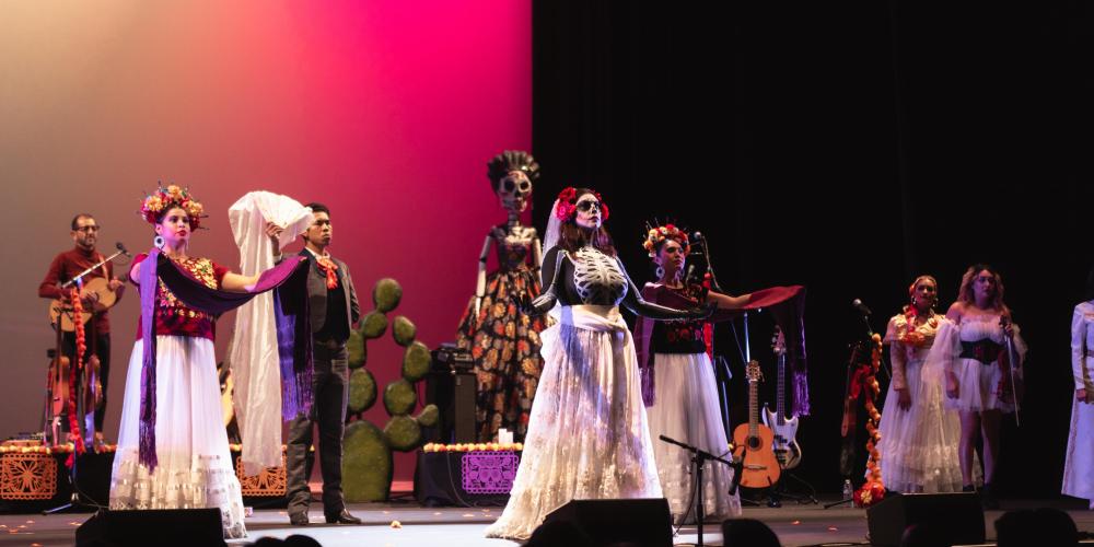 8 performers dressed in dia de los muertos themed attire  on stage with pink backlight 