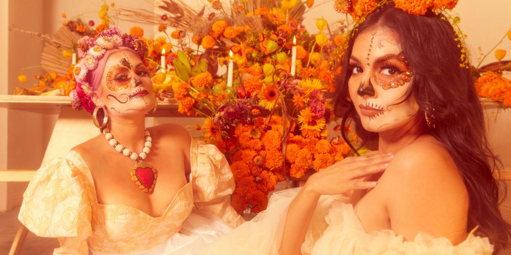 Two women pose in white dresses, orange flowers behind them and their faces painted like skeletons